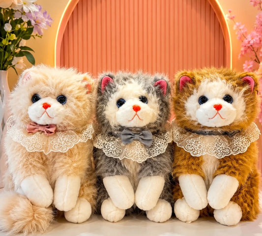 Three plush cat toys arranged side by side, each with long fluffy fur, white paws and faces, and lace necklaces with bows. The left toy is cream with a pink bow, the center toy is gray and white with a gray bow, and the right toy is golden brown with a dark bow.